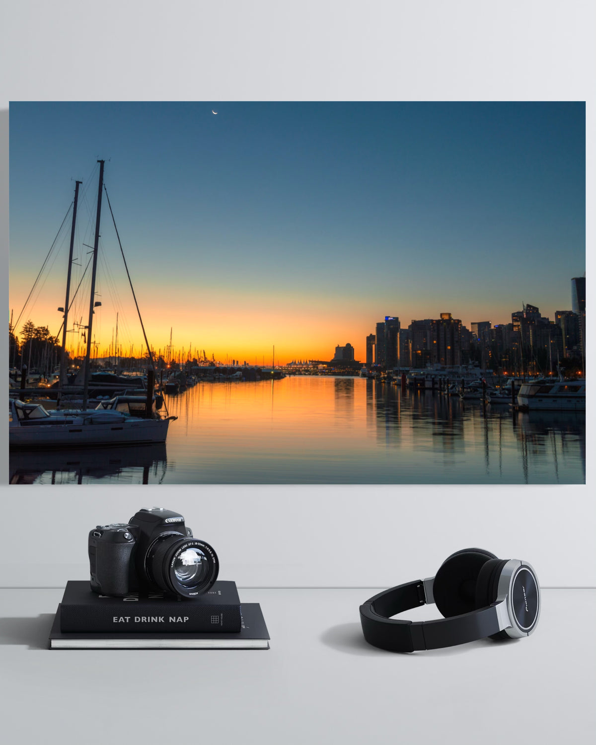 Close view of the Vancouver Sunrise print showing sailboat masts, a crescent moon and the city skyline at dawn, above a shelf with a camera and headphones