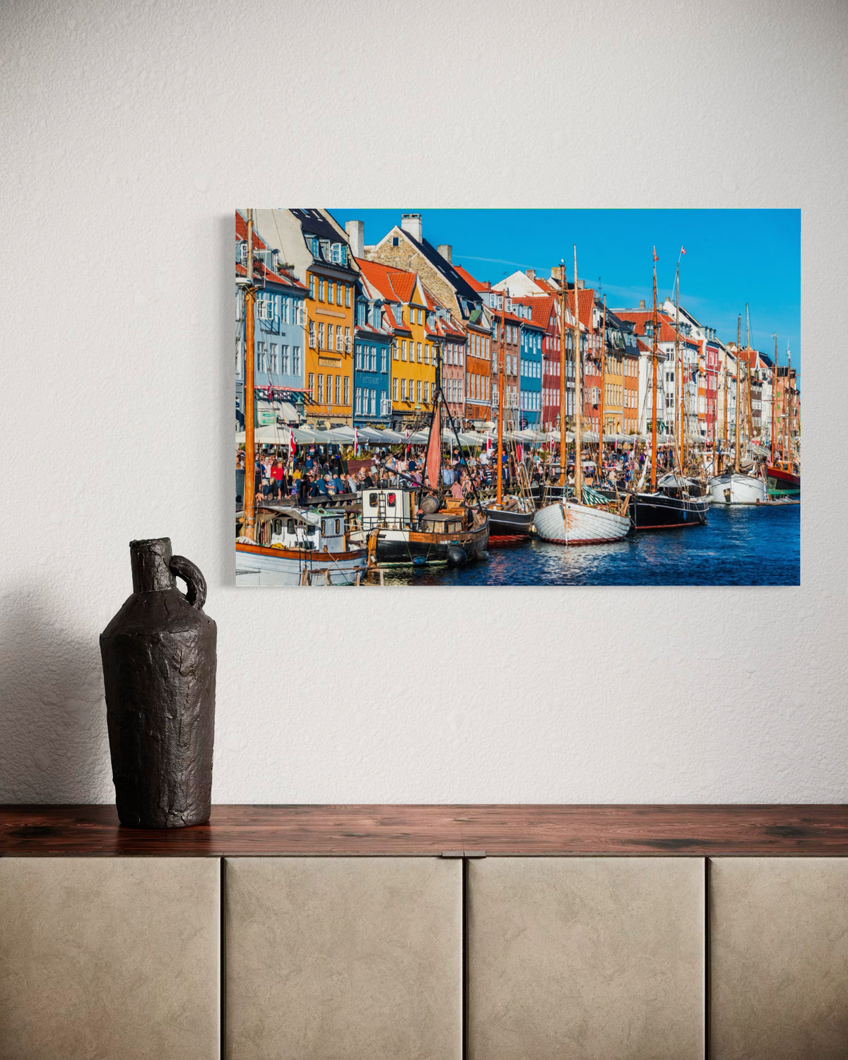 Nyhavn print on a textured white wall above a wooden sideboard with a dark ceramic jug