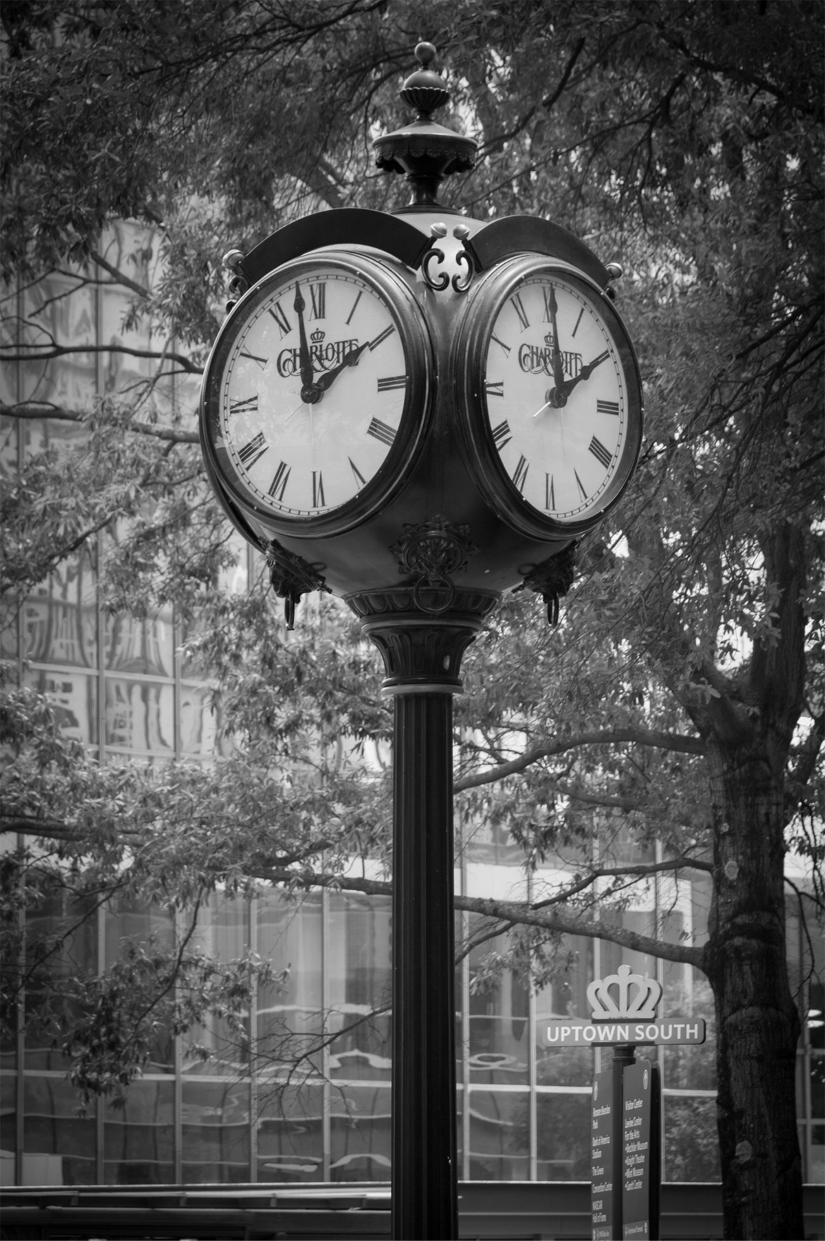 Ornate street clock with Roman numerals and the Charlotte name and crown on its faces, on a black post in uptown Charlotte, with trees, a glass office building and an Uptown South sign behind, black and white. The Clock fine art photography print