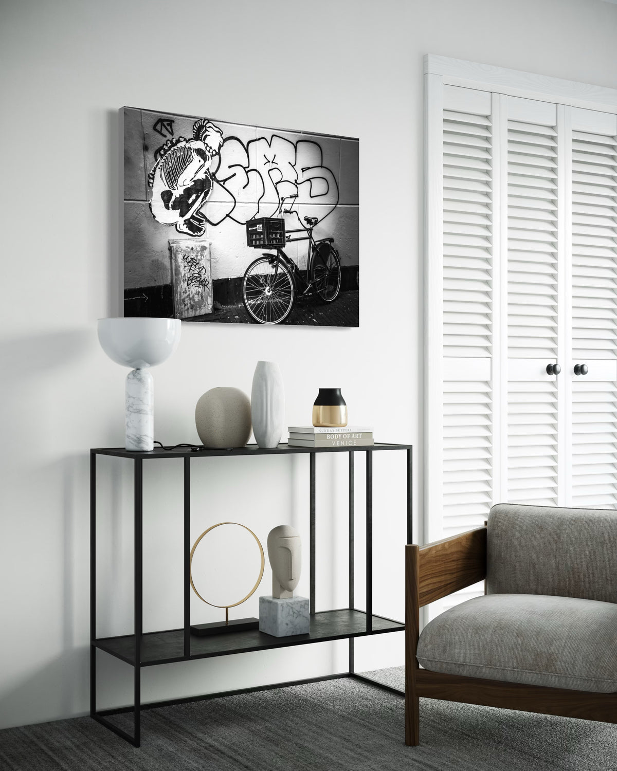 Street & Steel print above a black metal console with vases, books and a marble lamp, beside a grey armchair and white louvered doors